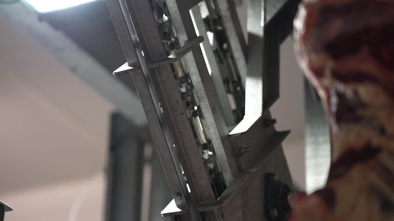 Primo piano di un nastro trasportatore a ganci in un impianto di lavorazione della carne, con binari metallici che si muovono su e giù