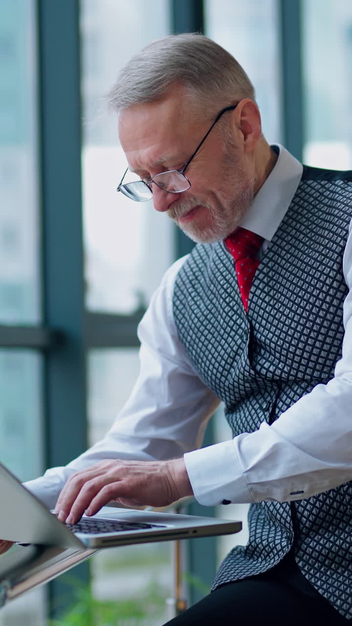 Mature businessman looking at laptop reading and printing improvements sitting near the window in his office. Panoramic city view background. Business video. Vertical video