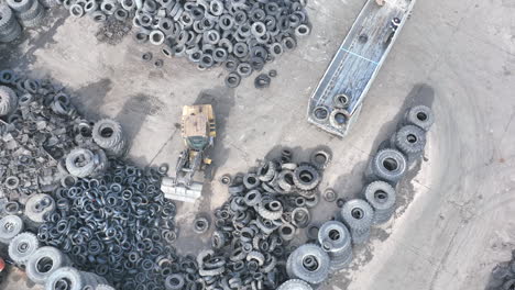 Toma-Aérea-De-Arriba-Hacia-Abajo-De-Un-Depósito-De-Chatarra-De-Reciclaje-De-Caucho,-Mientras-Un-Empleado-En-Un-Cargador-Frontal-Clasifica-Y-Apila-Los-Viejos-Neumáticos-Usados-Después-De-Que-Un-Camión-Haya-Descargado-Un-Gran-Remolque-Lleno-De-Material