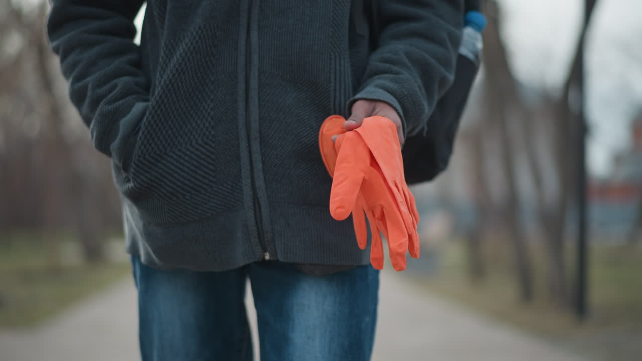 Closeup of young Indian man's torso outdoors holding orange gloves in right hand while other hand is in hoodie pocket, wearing jeans and dark sweater, standing on empty paved walkway in park