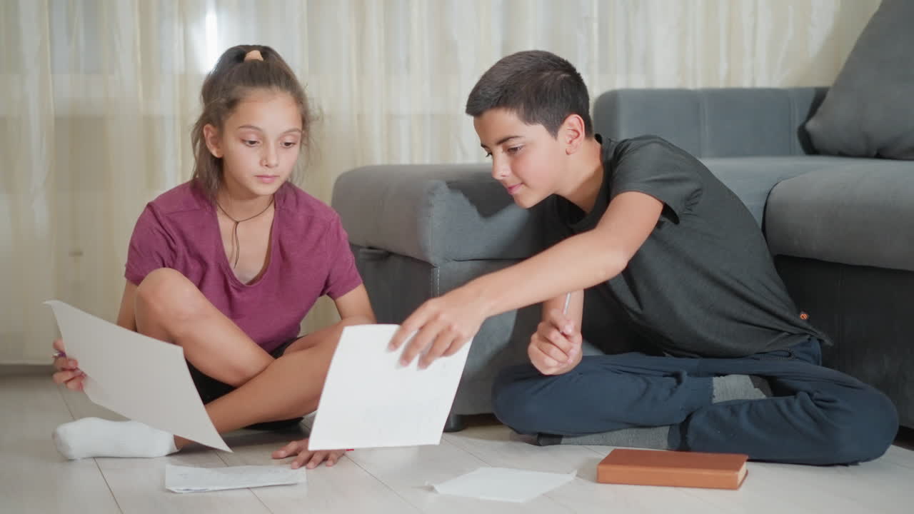 Amigos sentados en el suelo haciendo los deberes, compartiendo ideas y sonriendo mientras un chico asiente aprobando y sigue escribiendo cerca de un sofá gris en una habitación acogedora con luz natural brillante que entra por las cortinas.