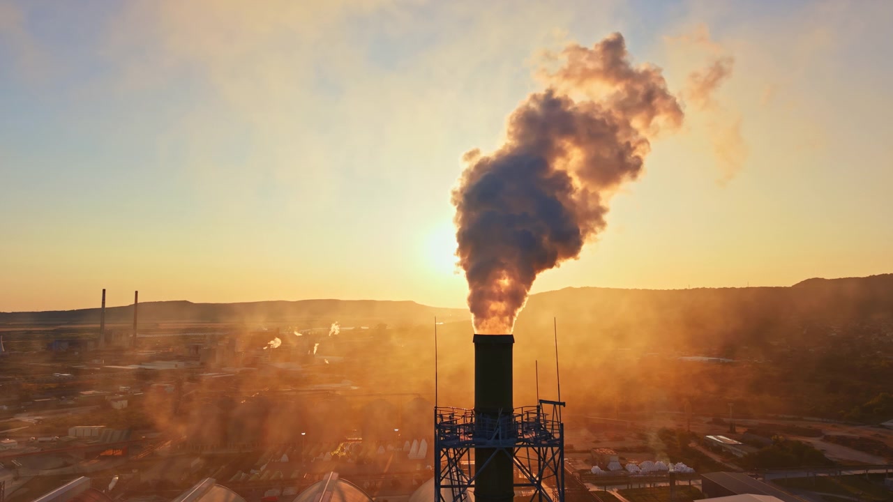 Fumo che sale da una ciminiera di fabbrica all'alba su un'area industriale circondata da colline.