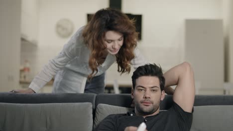 Happy-couple-smiling-in-living-room.-Concentrated-man-checking-tv-channels.