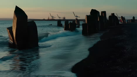 twilight waves on a dark sand beach with concrete sea defenses and distant port cranes