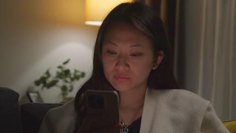 close up of woman spending evening at home sitting on sofa with mobile phone scrolling through internet or social media 3