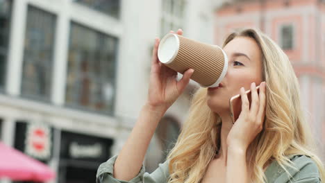 Close-up-view-of-Caucasian-blonde-woman-talking-on-the-smartphone-while-walking-down-the-street-and-drinking-coffee-to-go