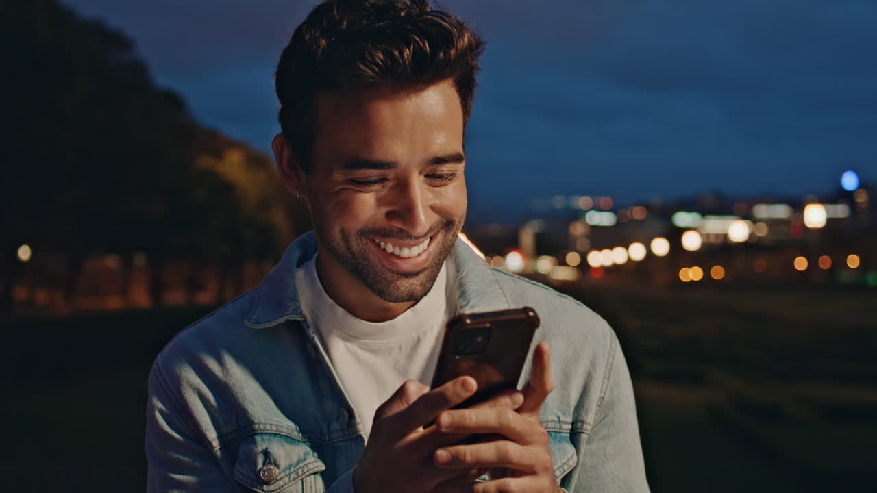 Uomo che ride risponde a un messaggio sullo smartphone mentre si rilassa in città di notte, primo piano.