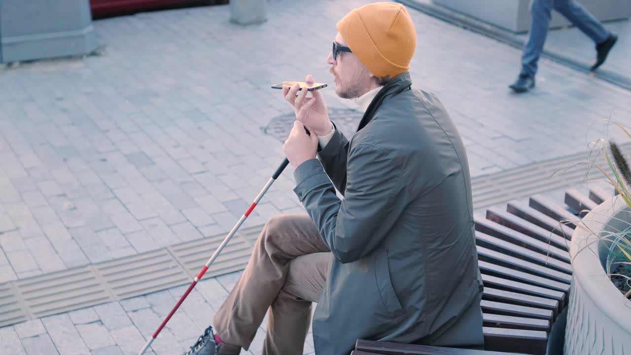 Young blind man with smartphone sitting on bench in park in city, calling