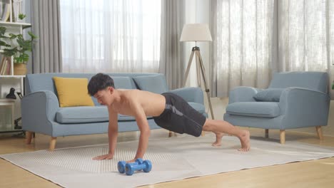 full body of asian teenager boy doing push ups on mat with dumbbells during the shirtless workout at home