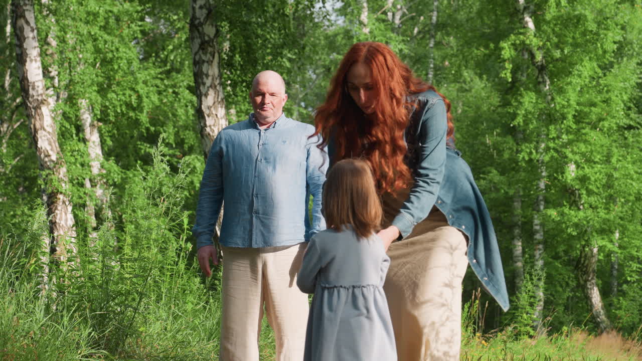 Little girl standing still refusing to join parents on forest path as mother gently moves closer, surrounded by lush green trees, expressing emotion, family connection, and caring outdoor moment