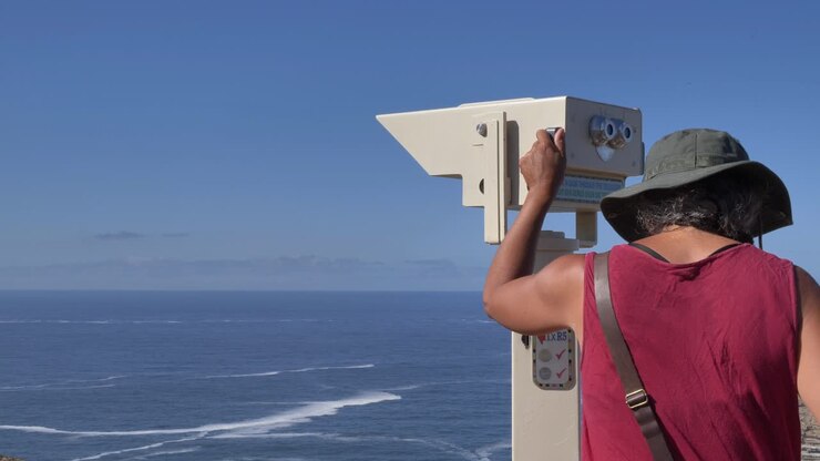 Una donna con un cappello e un vestito rosso usa un telescopio a gettoni per osservare l'ampio oceano blu e l'orizzonte lontano da Cape Point, Penisola del Capo, Sudafrica