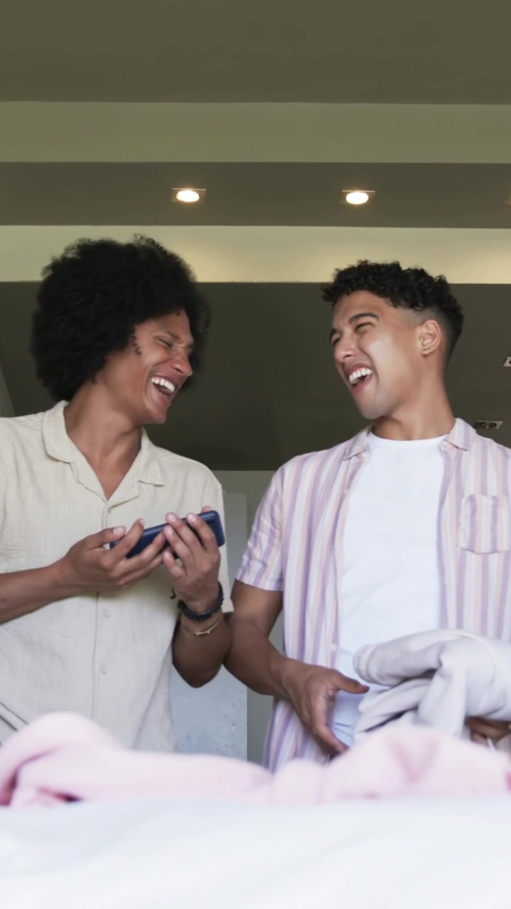 Vertical video: Showing phone, man in striped tee laughing while pal placing towels in box on bed