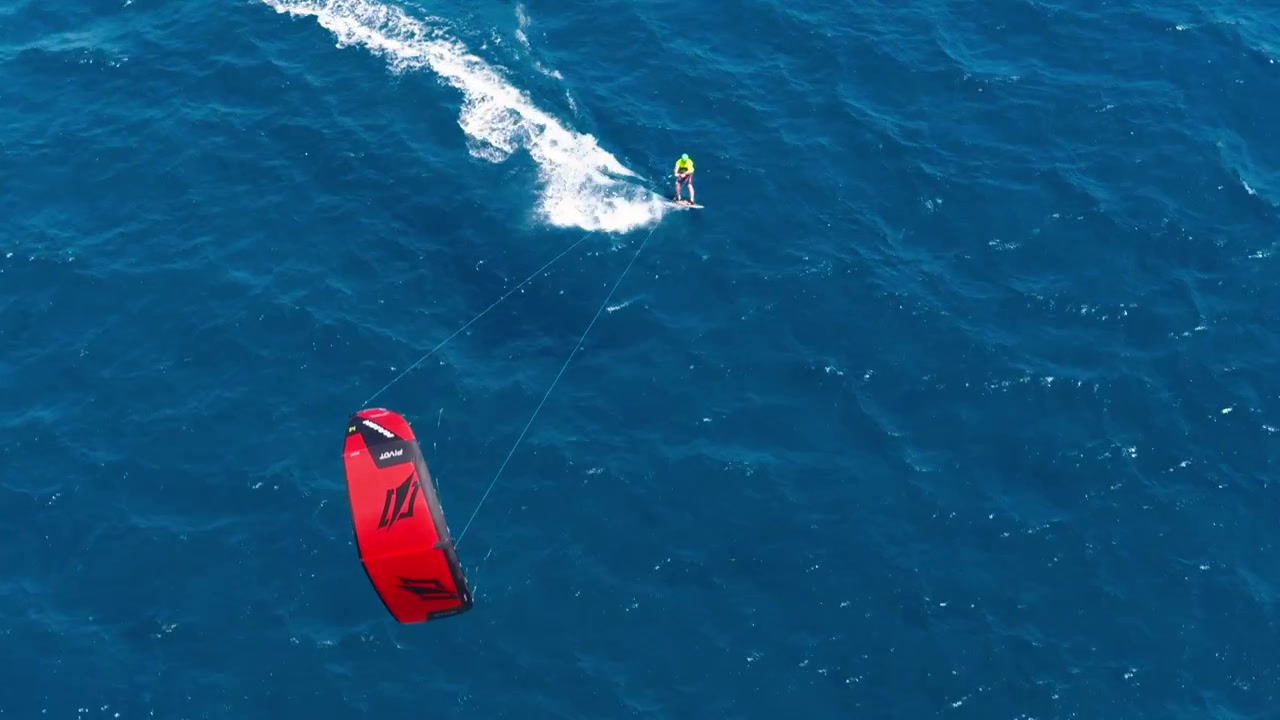 Et bredt luftfoto giver et fuldt overblik over et travlt kitesurf-sted med flere kitesurfere aktive over bugten og kystlinjen synlig i baggrunden, ved La Caletta på Sardinien, Italien.