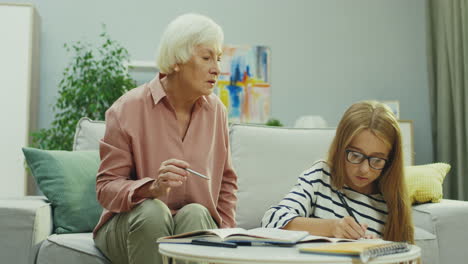 pretty blonde schoolgirl in glasses writing in a copybook as doing her homework and her grandmother helping her