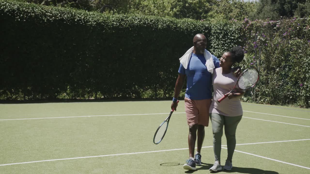 Felice coppia anziana afroamericana che cammina con racchette da tennis e si abbraccia al rallentatore