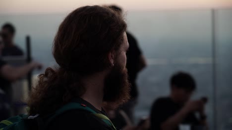 4k 24fps tracking shot where the camera follow a bearded man wearing a backpack turning around him on a skyscraper while he is contemplating the views from up there during sunset
