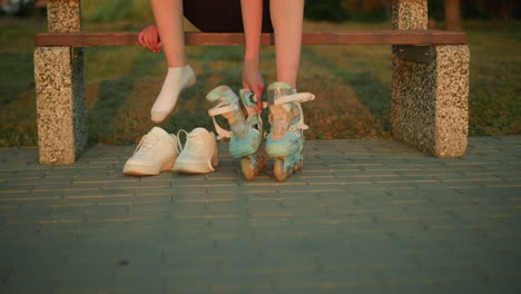 partial view of lady removing right leg roller skate while sitting on bench during sunset with two white sneakers placed beside her, with blurred background featuring greenery