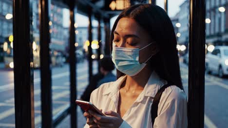 young woman wearing protective face mask standing at urban bus stop, using smartphone during evening city commute, representing pandemic lifestyle and digital communication