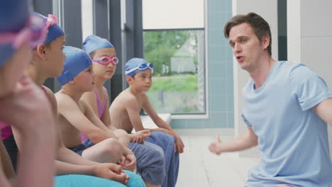 Male-Coach-Giving-Children-In-Swimming-Class-Briefing-As-They-Sit-On-Edge-Of-Indoor-Pool