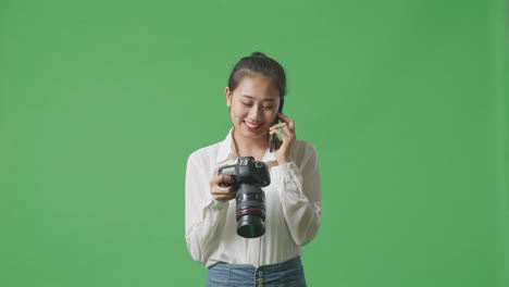 asian photographer holding a camera in her hands and talking on smartphone while standing on green screen background in the studio