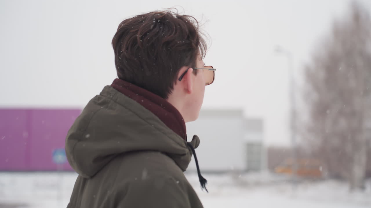 Teenager wearing winter jacket and sunglasses walks across snow covered parking lot toward parked car while looking around near urban buildings under falling snow on cold winter day