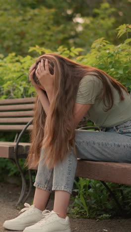 vista lateral de una mujer sentada en un banco de madera, las manos cubriendo suavemente su rostro en una postura reflexiva y sombría, su largo cabello cayendo en cascada por su hombro, rodeada de exuberante vegetación