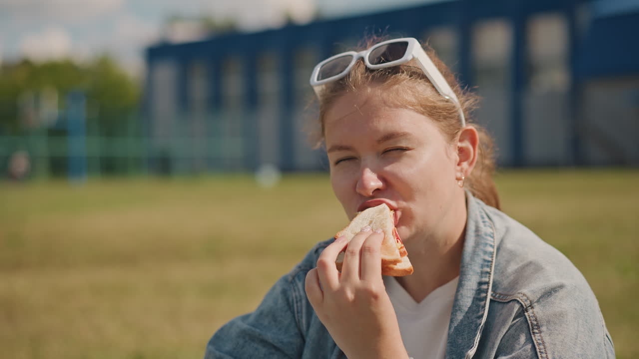 Giovane donna che si rilassa con del cibo all'aperto. Studentessa che pranza su un'area erbosa sotto un cielo luminoso. Donna con giacca di jeans che sorride mentre addenta un panino in un pomeriggio soleggiato all'aperto