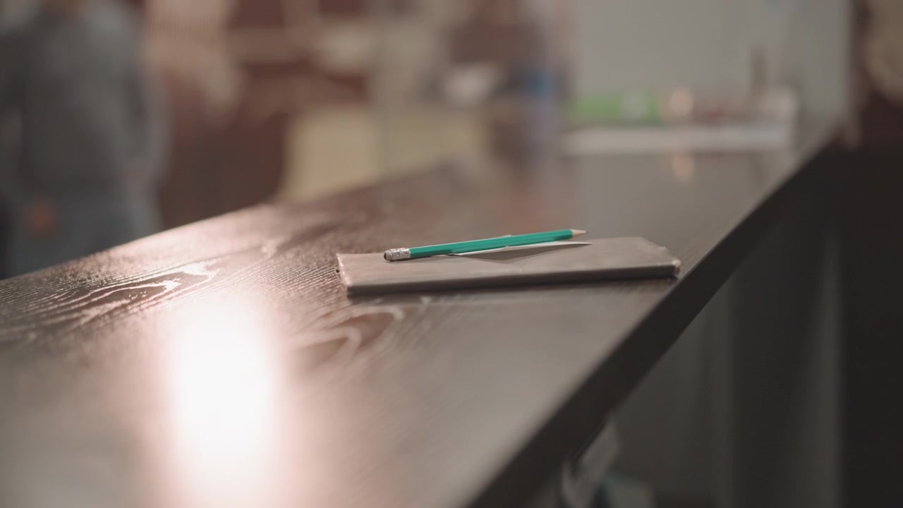 Tomt skrivebord med blyant og notesbog, varmt bokeh-lys på træoverflade i musikskolelobby, diskret refleksion, lærer efterlod hurtige noter før lektionen, rolig forberedende atmosfære