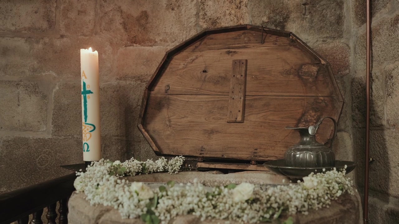 Cirio pascual encendido y flores blancas junto a una pila bautismal de piedra en una iglesia.