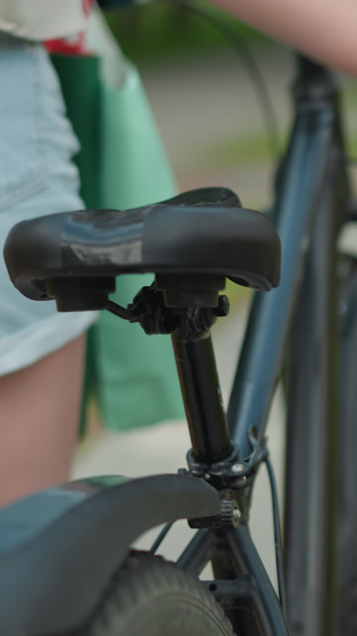primer plano de una mujer joven en pantalones cortos de jean y crop top caminando en su bicicleta, con un bolso negro a su lado y una vista parcial de la bolsa de menta en el frente, paseando por un camino pavimentado del parque rodeado de vegetación