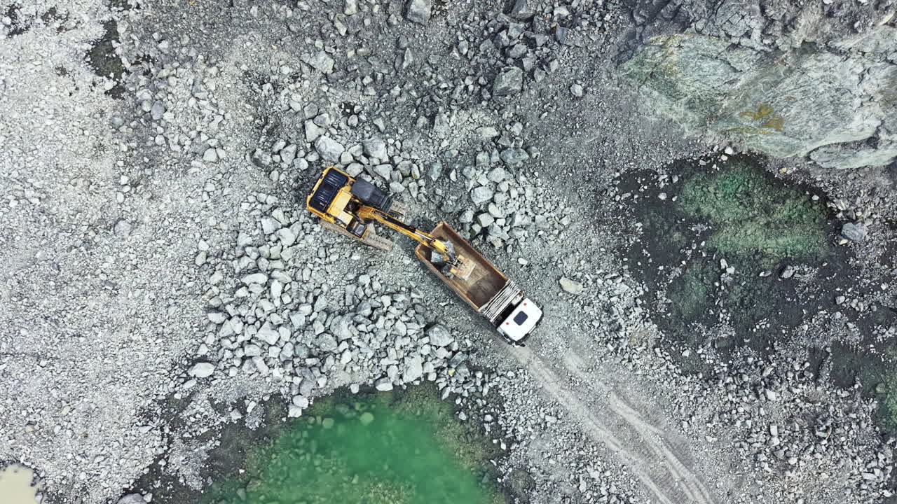 Gravemaskine og lastbil arbejder med at fjerne granitsten fra et stenbrud i et stenet terræn i dagtimerne.