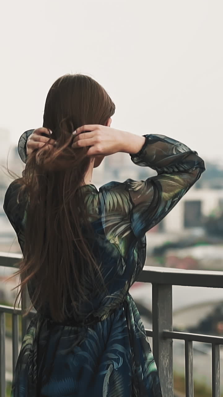 Graceful lady wearing stylish dress with chiffon decor adjusts hair waving in wind on viewing deck against large city slow motion backside view