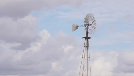 Wide-4K-shot-of-an-old-rustic-farm-windmill-blowing-and-spinning-in-the-wind-with-a-partly-cloudy-blue-sky-in-the-background