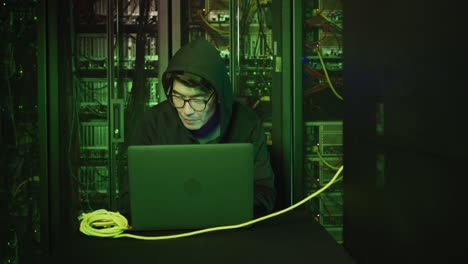 asian male hacker in hoodie using laptop by computer servers