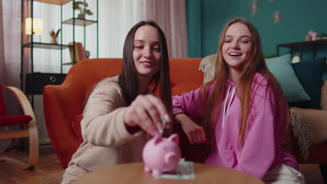 girls friends siblings sitting on floor and take turns dropping dollar banknote into piggy bank