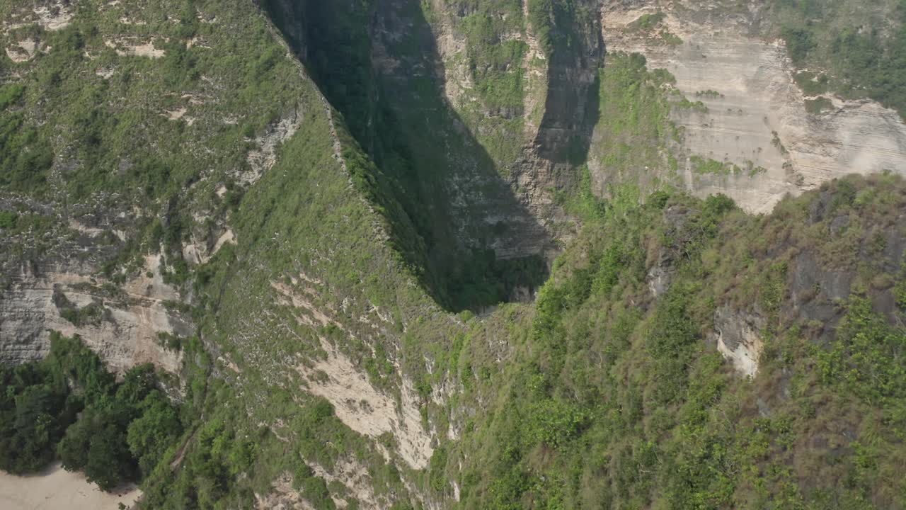 Aerial drone shot in the iconic Kelingking Beach, the 'T-Rex' shaped headland, and the towering limestone cliffs of Nusa Penida. Turquoise Indian Ocean crashes against the rocks and white-sand beach