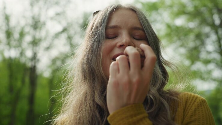 Woman with long hair wearing yellow sweater is outdoors holding something close to their face. background shows green trees and natural light suggesting scene takes place in nature or park