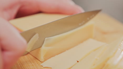 extreme close up of hand holding knife slicing through yellow dairy block on wooden cutting board, showing texture and precision of food preparation during home cooking in bright kitchen setting