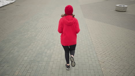 vista trasera de una mujer corriendo al aire libre en el pavimento entrelazado durante el invierno, el cabello que fluye mientras corre, el suelo cubierto de nieve y la olla de flores acentúan el ambiente urbano sereno