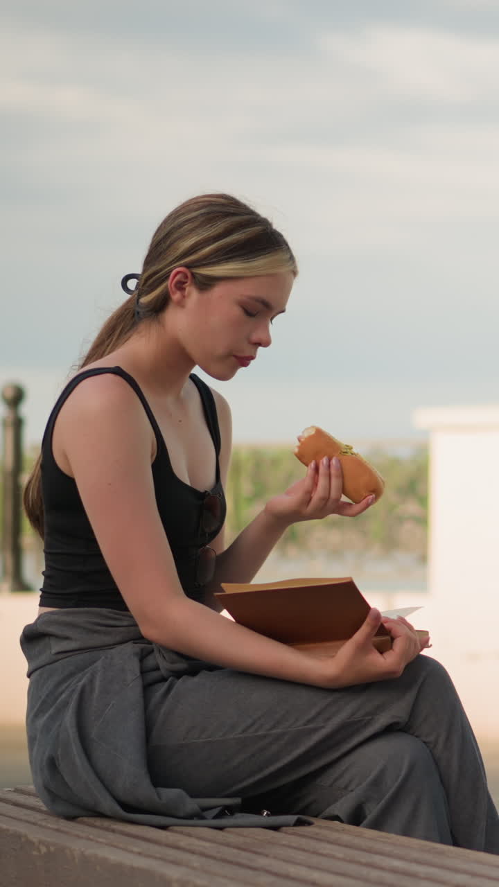 mujer en camiseta negra sentada al aire libre, comiendo una hamburguesa mientras lee un libro con una valla en el fondo, en un entorno al aire libre bajo un cielo brillante, disfrutando de un bocadillo casual y leyendo
