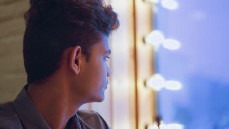 Young man sitting indoors near window, staring into distance with calm expression on face, surrounded by soft blue ambient light and bokeh glow, evoking mood of introspection, and peaceful contemplation