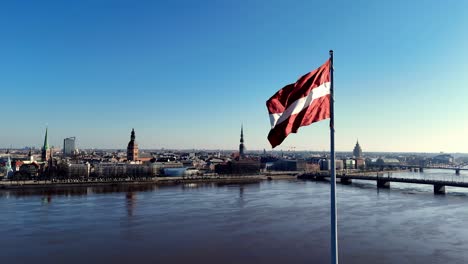 Lettische-Flagge-Weht-Im-Wind-Mit-Brücken-über-Den-Fluss-Daugava-Und-Der-Stadt-Riga-Im-Hintergrund-In-Lettland