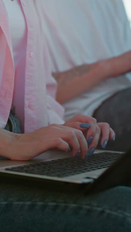 primer plano de una mujer joven con camiseta rosa y vaqueros escribiendo en una computadora portátil con uñas azules mientras está sentada al aire libre, al lado de otra persona también centrada en escribir en su dispositivo