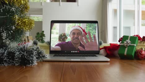 Smiling-biracial-man-with-santa-hat-waving-on-christmas-video-call-on-laptop