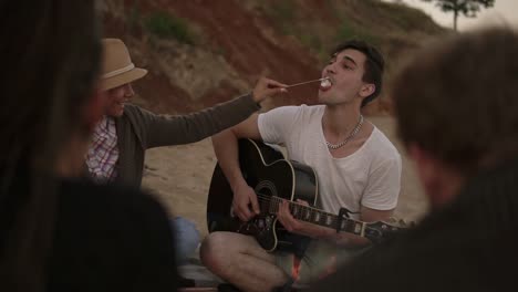 una giovane coppia romantica seduta accanto al fuoco sulla spiaggia insieme agli amici. l'uomo suona la chitarra e la donna dà