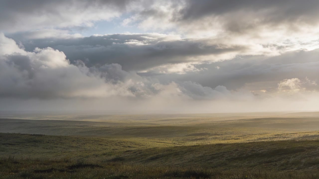Banchi di nuvole che si spostano rivelando fasci di luce solare, una pianura erbosa aperta che riceve i raggi, con la nebbia che si accumula.