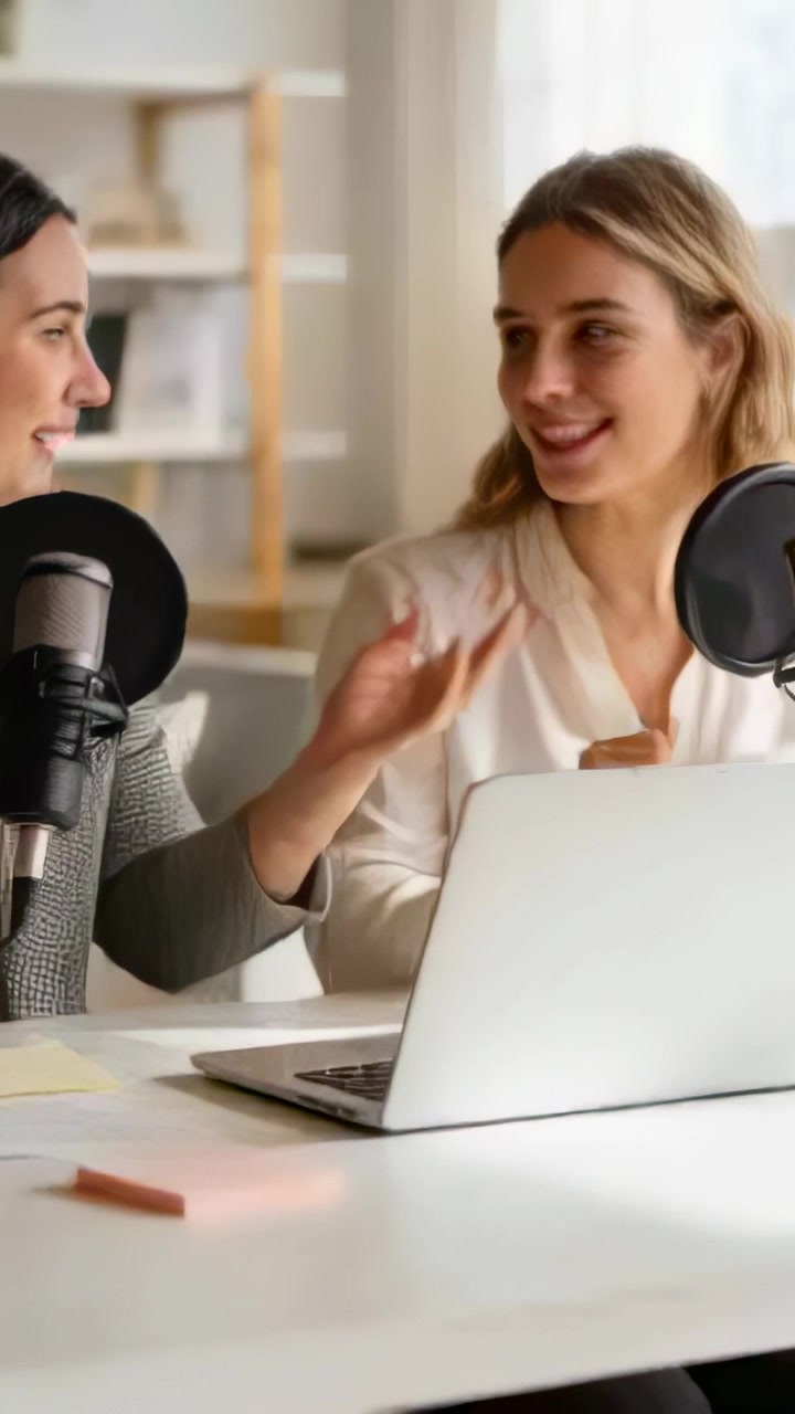 Video vertical: Dos presentadores con blazer y blusa revisan un portátil, preparando un episodio en casa con micrófonos.