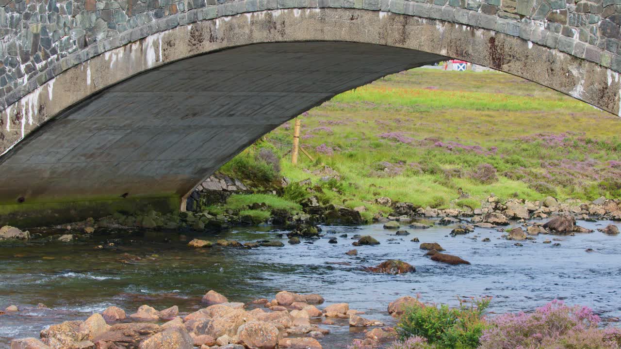 Wide shot of stone bridge spanning flowing stream, purple heather, and moorland under daylight