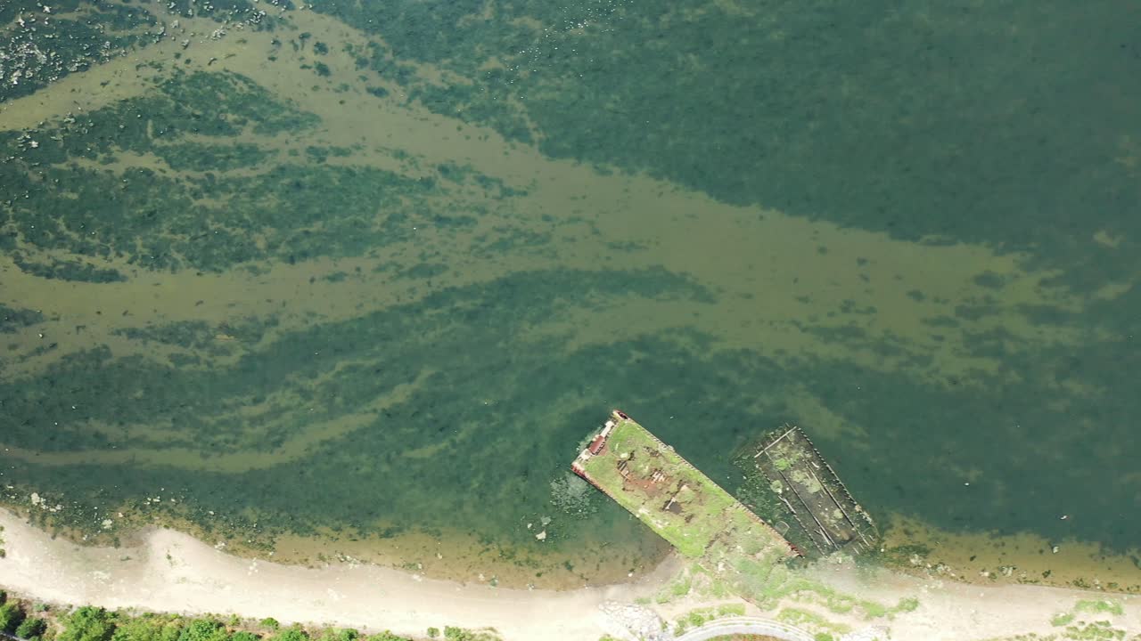 A drone shot over Gravesend Bay in Brooklyn, NY. It is a sunny day and the water is calm and reflective. There are green trees along the shore and algae on the water. The camera trucks to the right