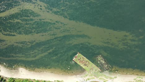 a drone shot over gravesend bay in brooklyn, ny. it is a sunny day and the water is calm and reflective. there are green trees along the shore and algae on the water. the camera trucks to the right